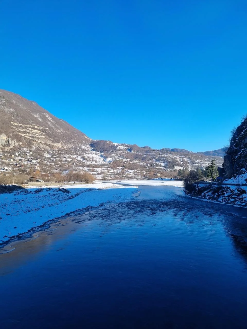 Partly frozen river mirroring blue sky, between snow covered mountains